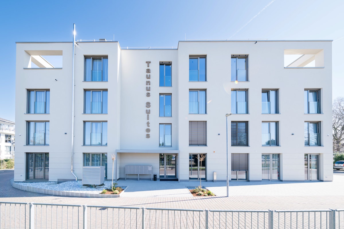 The modern, three-story building features a sleek exterior with large windows, providing ample natural light. The façade is enhanced by the name 'Taunus Suites' prominently displayed. A landscaped area with minimal greenery is visible at the entrance, offering a welcoming approach.
