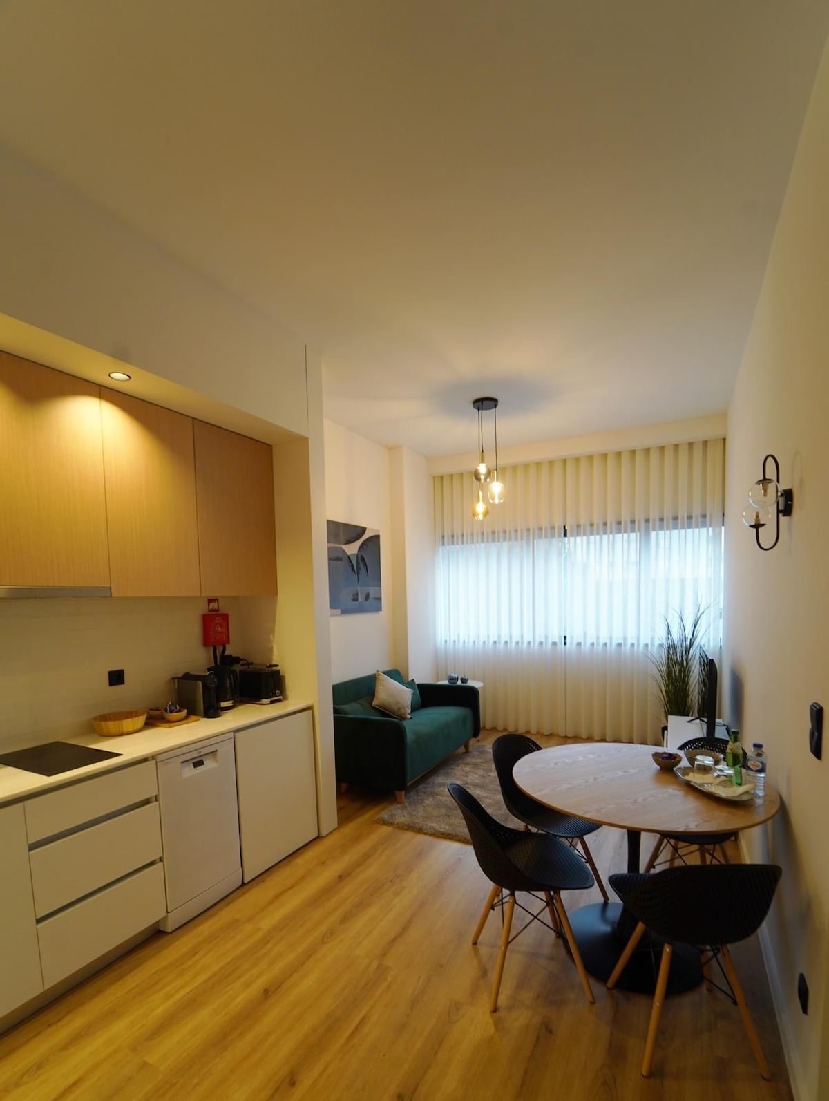 A compact kitchenette is seen, featuring light wood cabinetry and modern appliances, including a refrigerator and coffee maker. Adjacent to it, a small dining table with black chairs is positioned, illuminated by natural light from a window adorned with sheer vertical blinds.