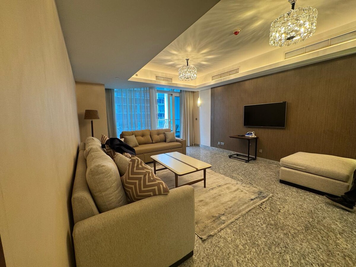 A spacious living area features a comfortable seating arrangement with neutral-toned sofas and decorative cushions. Large windows allow natural light to fill the space, while two elegant chandeliers provide soft illumination. A flat-screen television is mounted on the wall, complemented by a minimalistic coffee table.