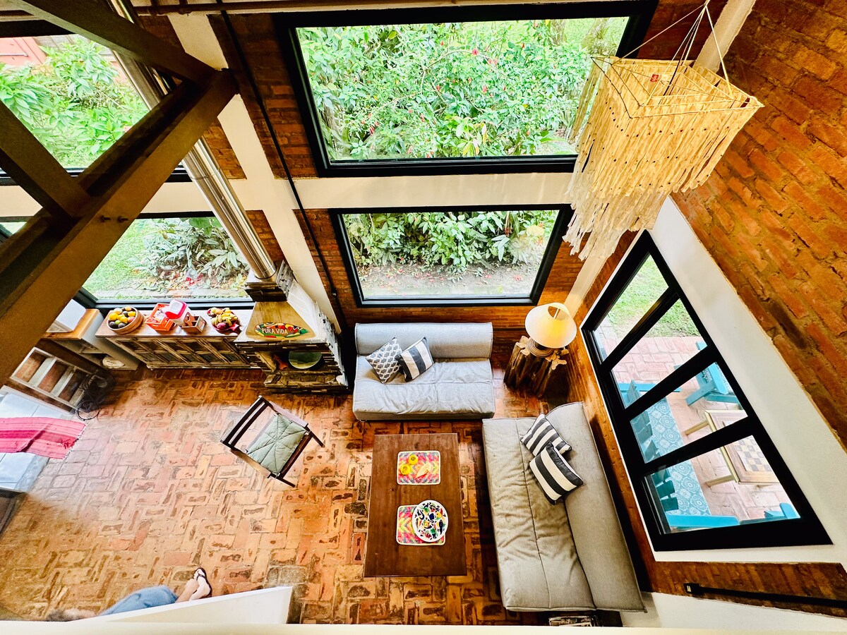 An inviting living area features large windows that fill the space with natural light, offering views of lush greenery. Two sofas are positioned around a coffee table with vibrant decor. A unique light fixture hangs above, enhancing the rustic charm of the wooden floors.