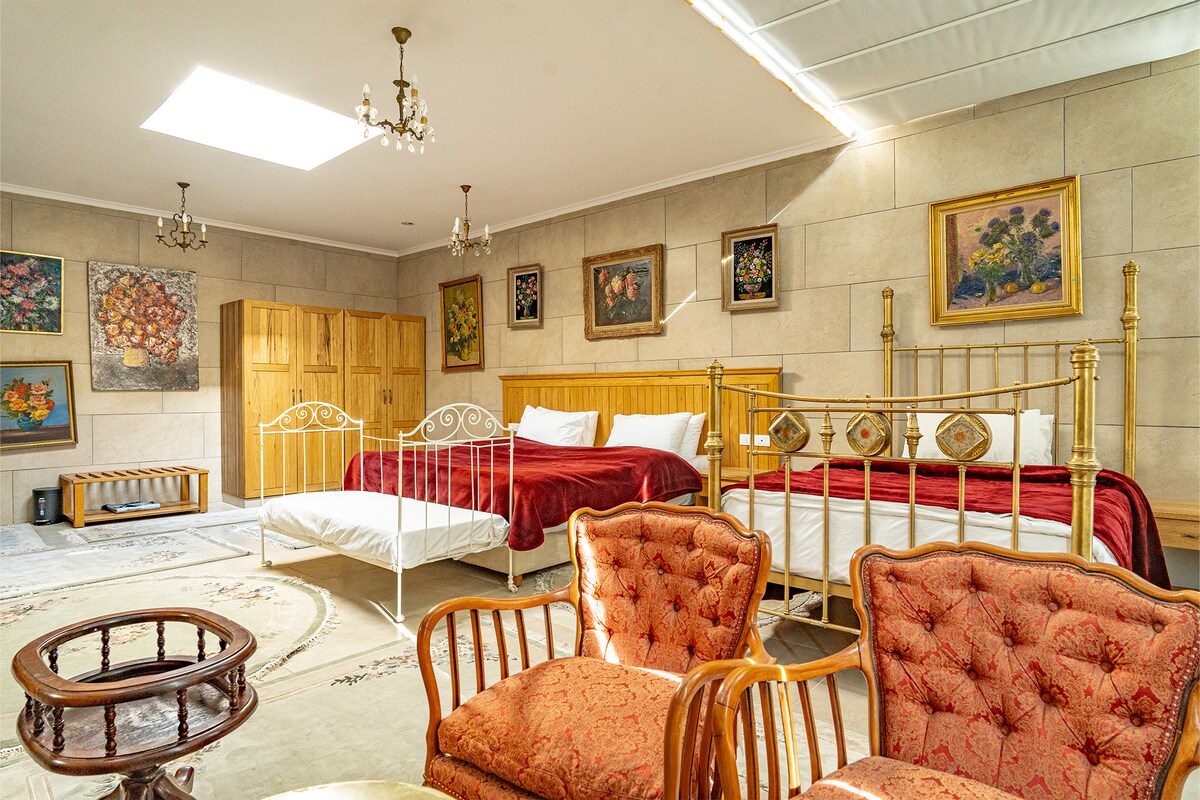 A spacious room is highlighted by two large beds with decorative headboards, complemented by a single bed nearby. Elegant artwork adorns the walls, and a traditional round table with chairs offers a cozy seating area. Natural light floods the space through a skylight.