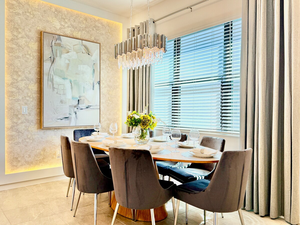 A modern dining area features a large, round wooden table surrounded by six upholstered chairs. A chandelier hangs above, illuminating the space. Sunlight filters through large windows adorned with elegant curtains, casting a warm glow on the decorative wall art.
