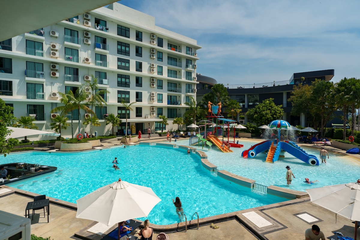 A large outdoor pool is surrounded by palm trees and lounge chairs with umbrellas. Water slides are visible in one corner of the pool area. Guests can be seen enjoying the water, while the hotel building features multiple balconies overlooking the poolside.