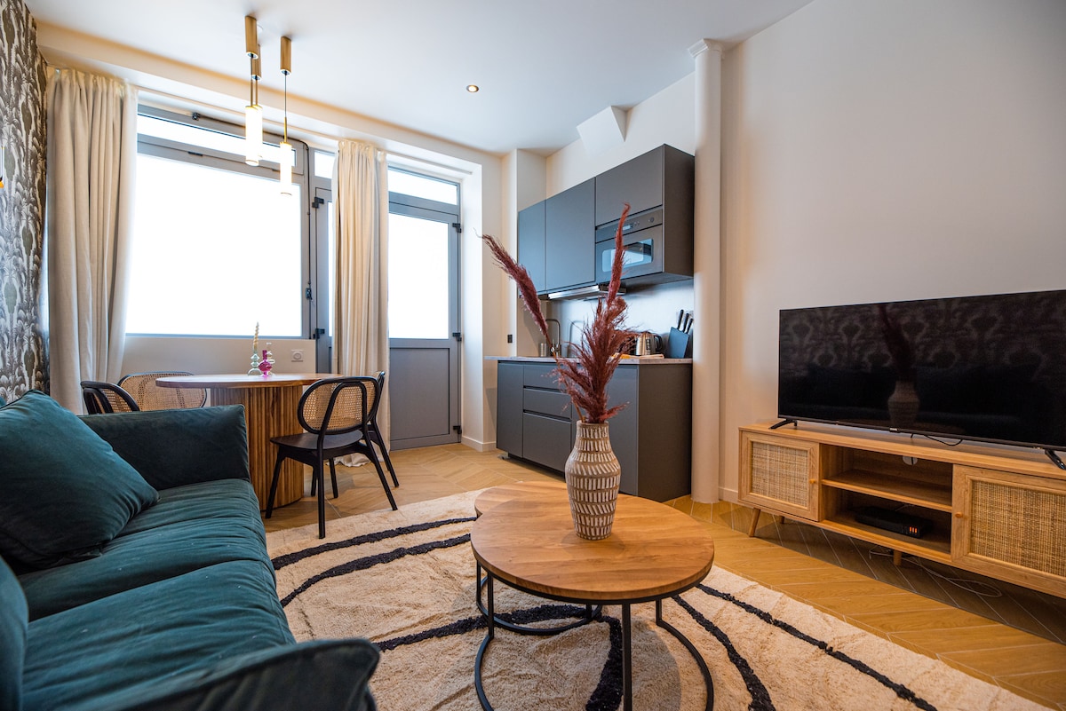 A modern living area features a plush green sofa and a round wooden coffee table. A flat-screen TV is positioned on a unit with woven accents. Natural light brightens the space through large windows adorned with light curtains, and a dining table set for two is nearby.