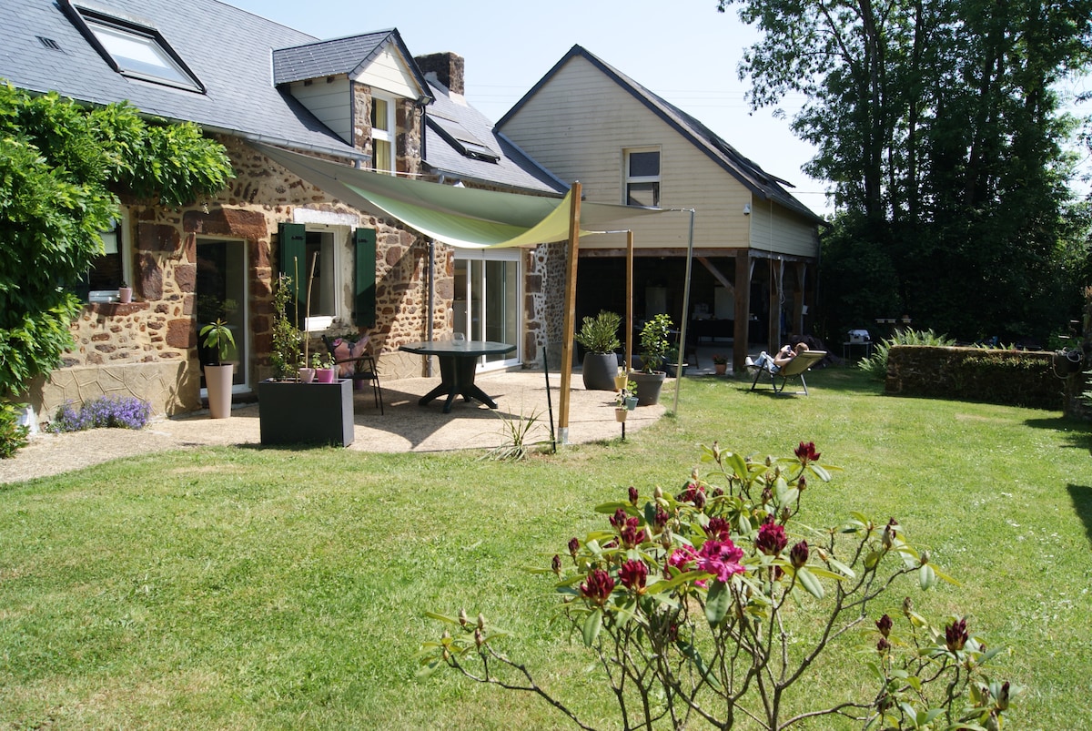 A spacious garden area is presented, featuring a well-maintained lawn and flowering shrubs. A stone building with window shutters is visible, complemented by a shaded patio space with outdoor seating. A secondary structure is observed in the background, providing additional shaded relaxation areas.