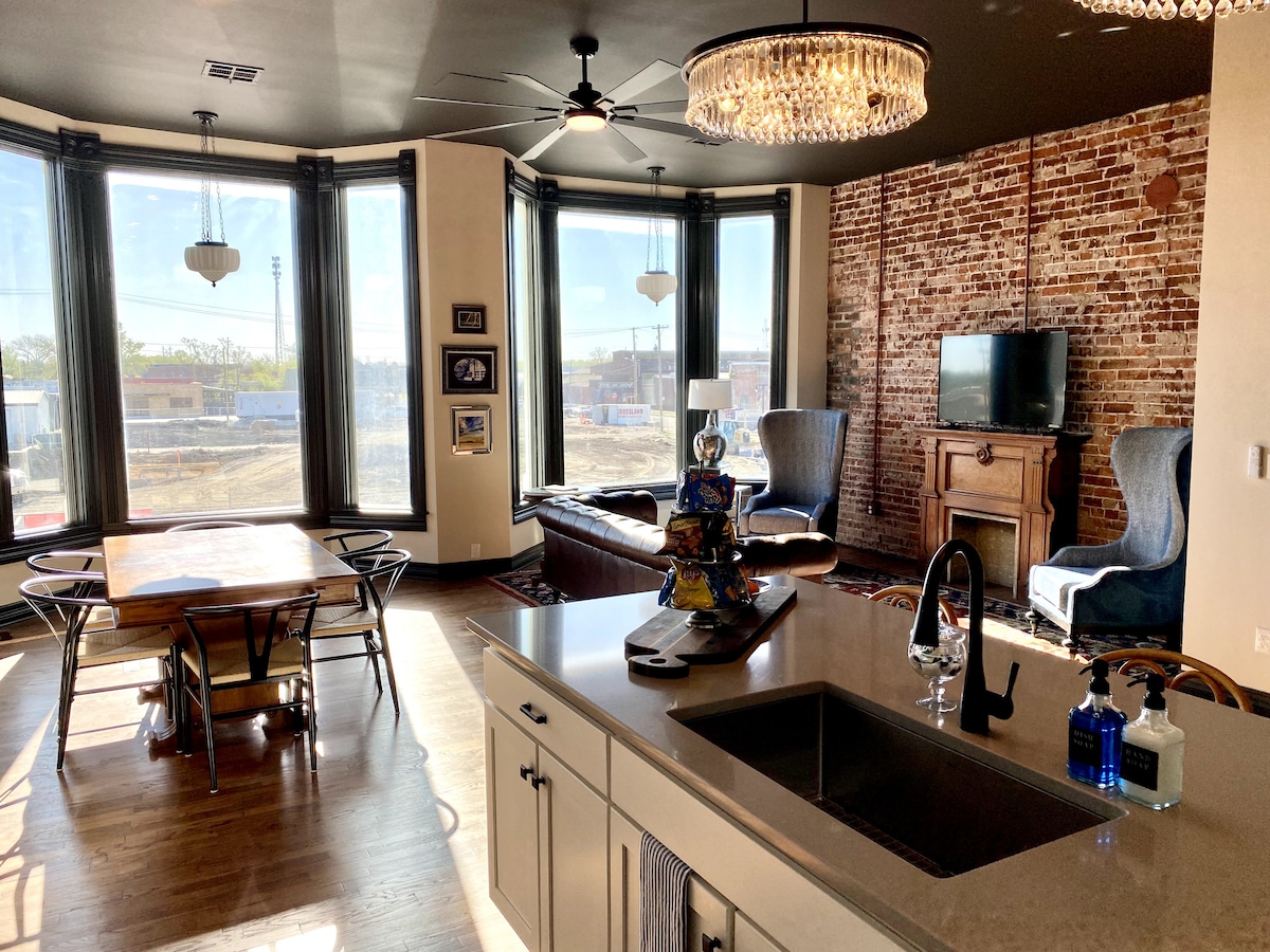 A modern living space features large bay windows that flood the area with natural light. A comfortable seating arrangement includes two armchairs and a sofa. A dining table is set nearby, and an open kitchen showcases sleek countertops and contemporary fixtures.