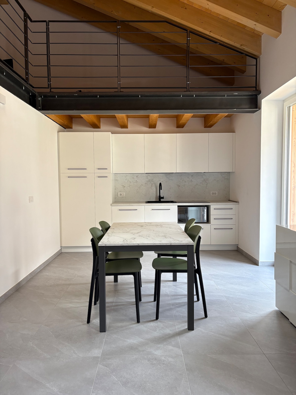 A bright kitchen area features minimalist cabinetry with a marble countertop. A small dining table with four green chairs is positioned in the center. Natural light enters through a large window, and a staircase leads to an upper loft space.