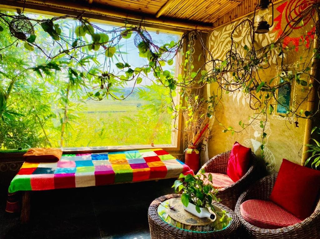 A charming seating area is presented with a colorful patterned blanket draped over a low bed. Two woven chairs with red cushions flank a small round table adorned with a potted plant. Natural light filters in through large windows, enhancing the view of the lush surroundings.