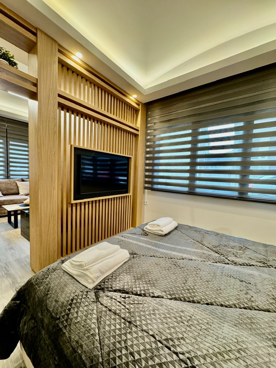 A cozy sleeping area features a grey textured blanket draped over a spacious bed. A mounted television is positioned on the textured wooden wall. Large windows with layered shades allow soft light to filter into the room, enhancing the modern design.