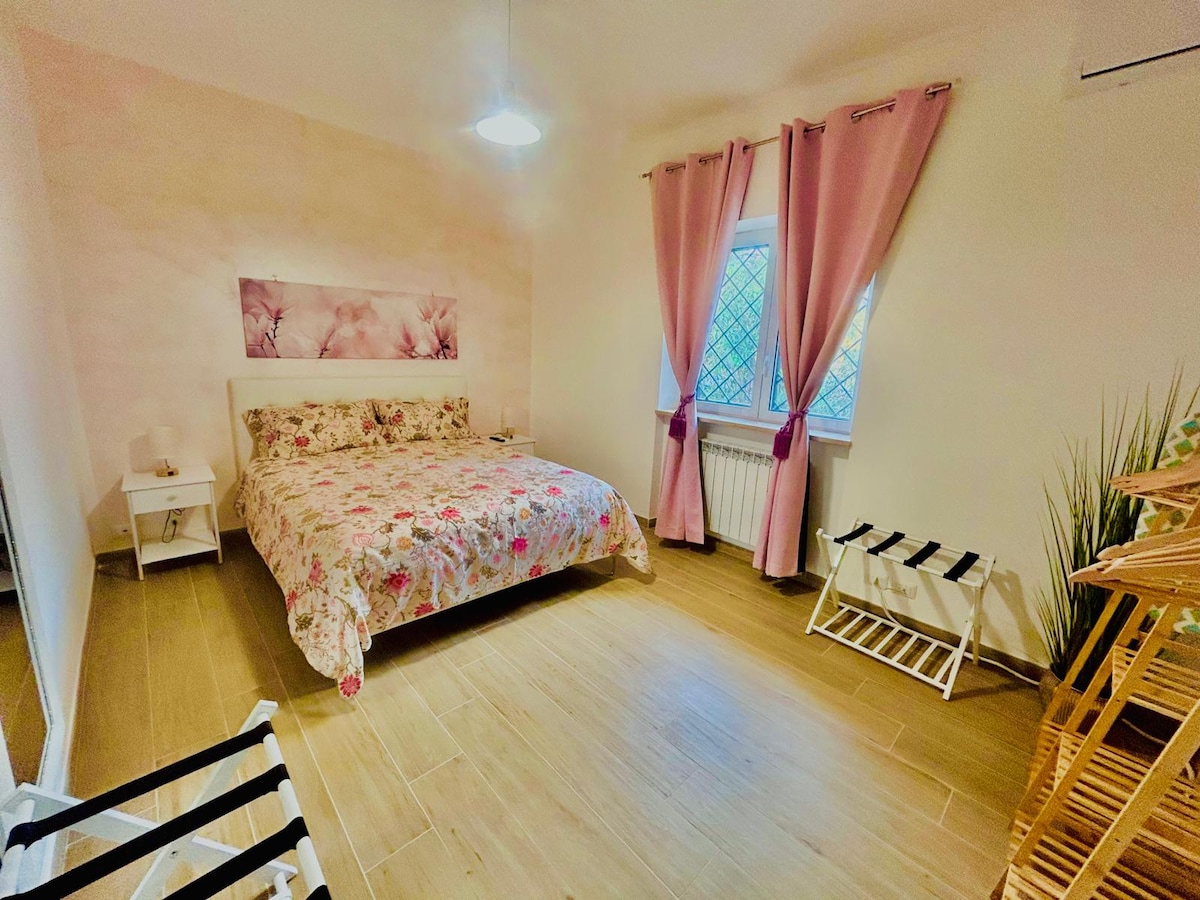 A bright bedroom features a spacious bed with floral bedding. A large window allows natural light to illuminate the space, complemented by soft pink curtains. Two bedside tables are positioned beside the bed, while additional folding chairs are arranged at the side.