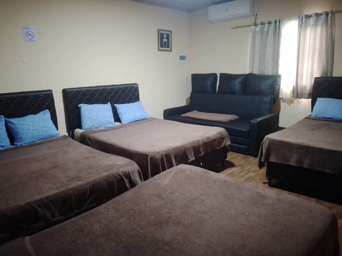 The sleeping area includes four spacious beds, each adorned with soft brown blankets and blue accent pillows. A dark sofa is positioned against the wall, providing additional seating. Natural light enters through a partially covered window, illuminating the cozy room.