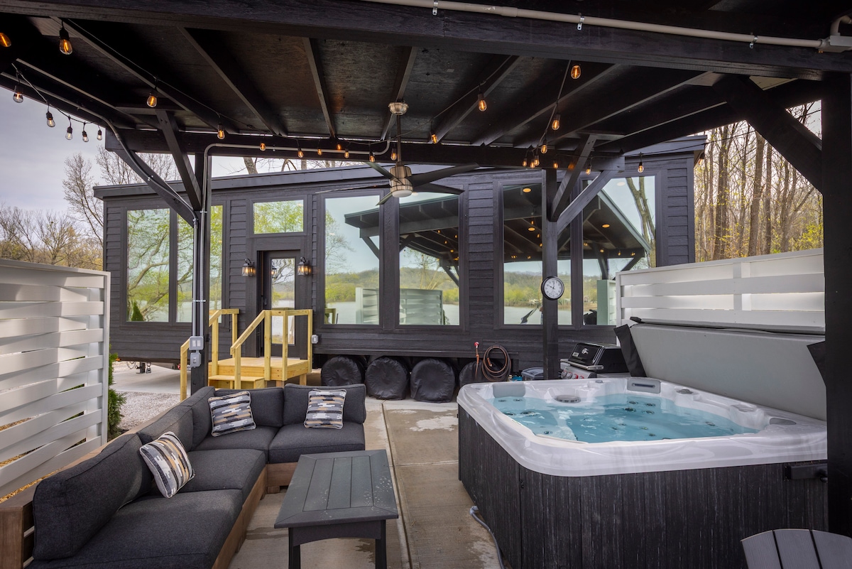 An inviting outdoor space features a covered patio with a large hot tub equipped with numerous jets. Comfortable seating is arranged around a dark table, and large windows reflect the surrounding nature, enhancing the serene ambiance of the tiny home.