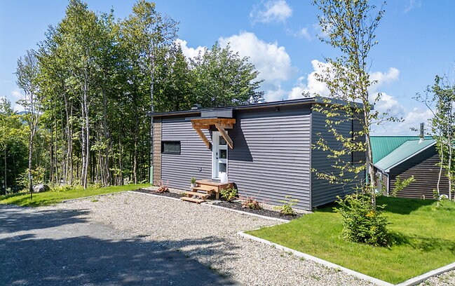 A modern chalet is set within a natural landscape, featuring a gray exterior complemented by wooden accents at the entrance. A gravel path leads to the front door, surrounded by greenery and young trees, providing a harmonious connection to the outdoors.