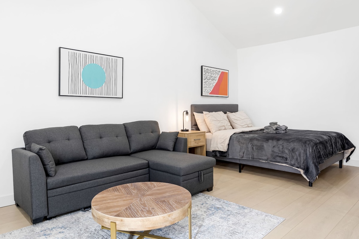 A modern studio space is highlighted with a cozy grey sectional sofa positioned beside a round wooden coffee table. A plush, neatly made bed with soft linens occupies the background. Minimalist artwork adorns the walls, enhancing the contemporary feel of the room.