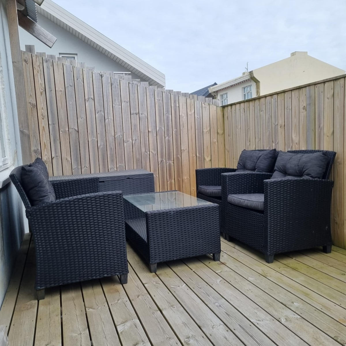 A wooden deck area is furnished with four dark wicker chairs featuring plush gray cushions and a matching coffee table with a glass top. The space is enclosed by a wooden fence, providing privacy.