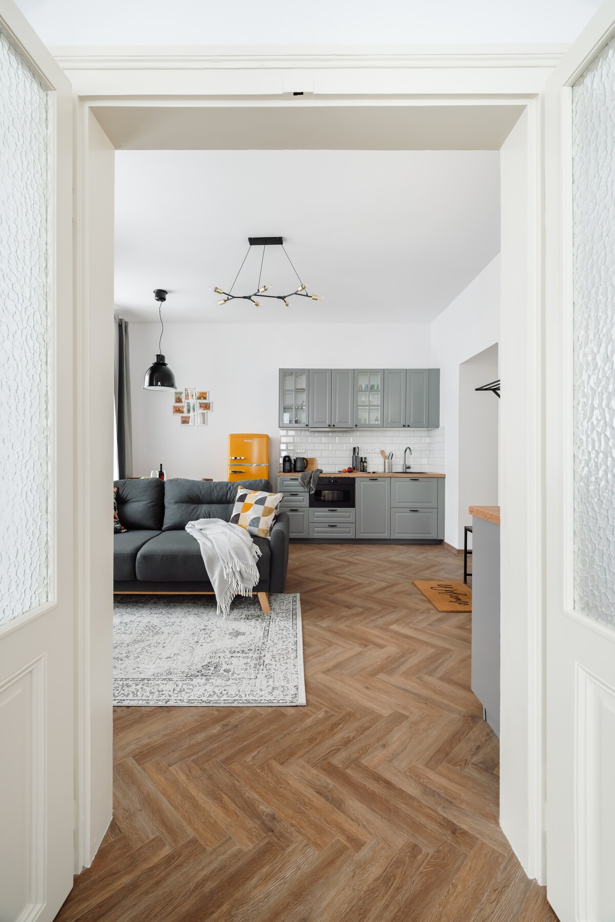 A view from an entryway reveals a modern living area featuring a gray sofa adorned with yellow and white pillows. A stylish kitchen with gray cabinetry and essential appliances is visible in the background, complemented by a patterned area rug on the warm wood floor.