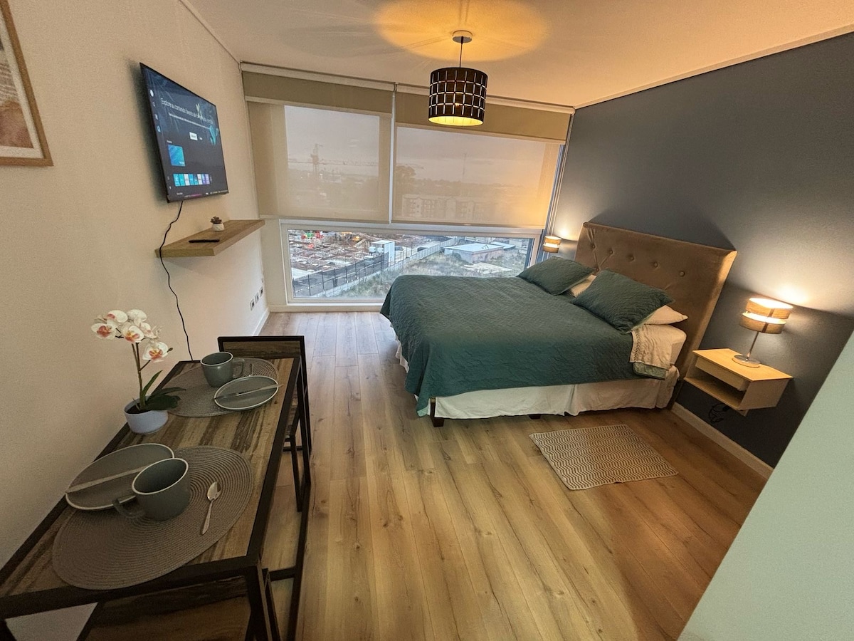 A modern studio room is presented, featuring a queen bed with teal bedding and accent pillows. A flat-screen TV is mounted on the wall, and a small dining table is set with tableware. Soft light is provided by bedside lamps, enhancing the airy atmosphere.
