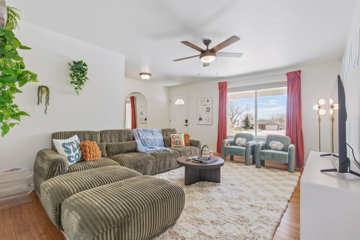 A spacious living room features a modular couch in muted tones and two stylish Cloud chairs. A ceiling fan is positioned above, and a sliding door allows natural light to illuminate the area. Soft, textured carpeting adds comfort to the space.
