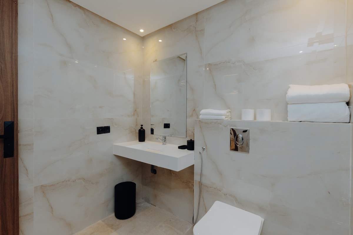 A modern bathroom is highlighted by sleek marble walls and a spacious, minimalist countertop. Soft, white towels are neatly arranged beside a mirror. Natural light enhances the clean, airy atmosphere, complemented by simple, stylish fixtures.