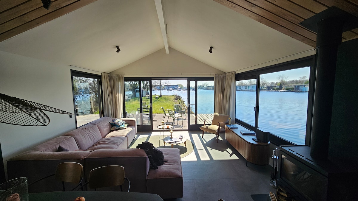 A spacious living area features large glass windows that provide unobstructed views of the water and garden. A comfortable sofa and wood accents create a cozy environment, while outdoor seating is visible on the patio. Natural light fills the space, enhancing the serene atmosphere.