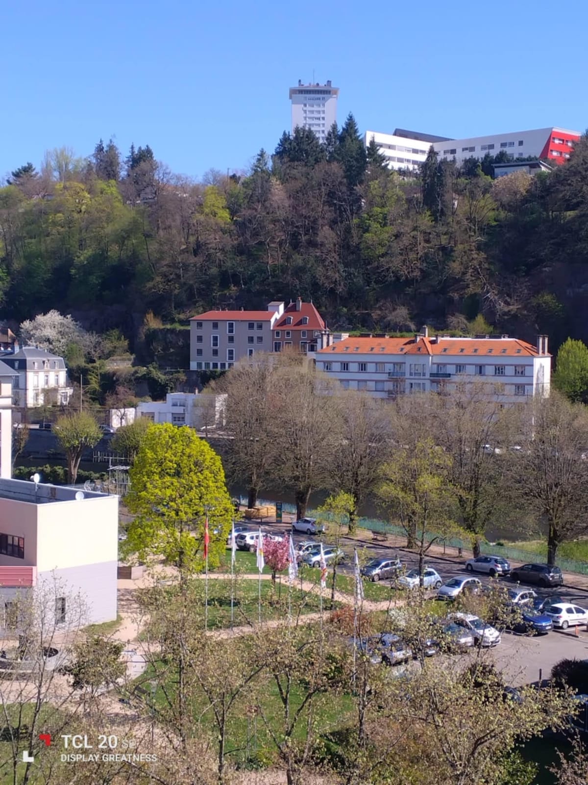 A view of a lush green landscape featuring a mix of trees and buildings. The foreground shows a cluster of cars parked along a road, while the backdrop includes charming structures nestled against a hillside. The sky is clear, enhancing the scene's brightness.