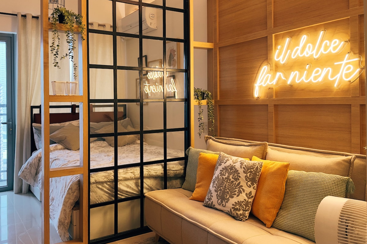 A cozy living area features a convertible sofa adorned with colorful cushions. A glass partition separates the space from the bedroom, where a queen-size bed is visible. Warm lighting enhances the ambiance, highlighted by neon text on the wall.