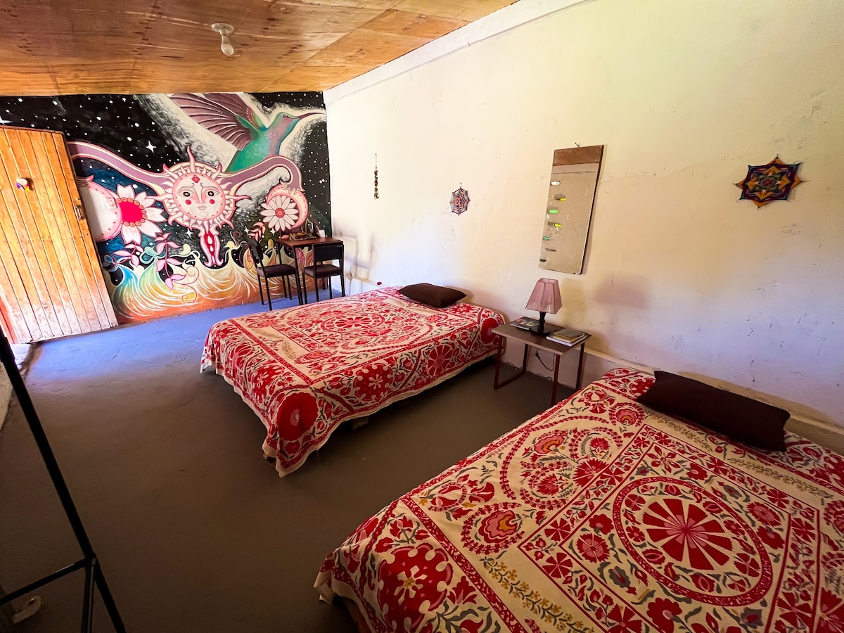A sleeping area features two spacious beds adorned with intricate red and white bedcovers. The colorful mural of nature and celestial themes enhances the room's ambiance. A small table and lamp are positioned against the wall, contributing to the overall welcoming atmosphere of the space.