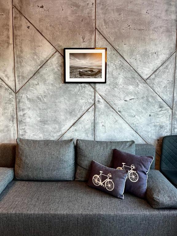 A cozy seating area features a gray sofa with plush cushions adorned with bicycle motifs. A framed picture hangs above the sofa, depicting a serene landscape, while the textured concrete wall adds a modern touch to the ambiance.