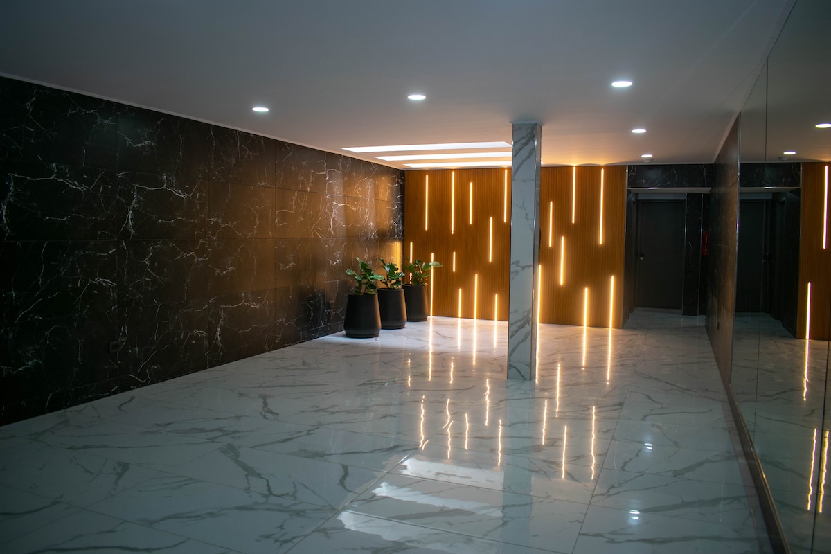 A modern entryway is displayed, featuring sleek black marble accent walls. A glossy white marble floor reflects soft lighting from vertical strip lights and recessed ceiling lights. Green plants are placed in simple black planters, adding a touch of nature to the contemporary design.