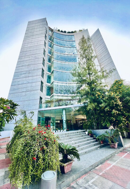 A modern multi-story building is prominently featured, showcasing large glass windows that reflect the surrounding greenery. The entrance is framed by potted plants and a set of stone steps leading to the main door. Lush trees are visible at the top of the structure.