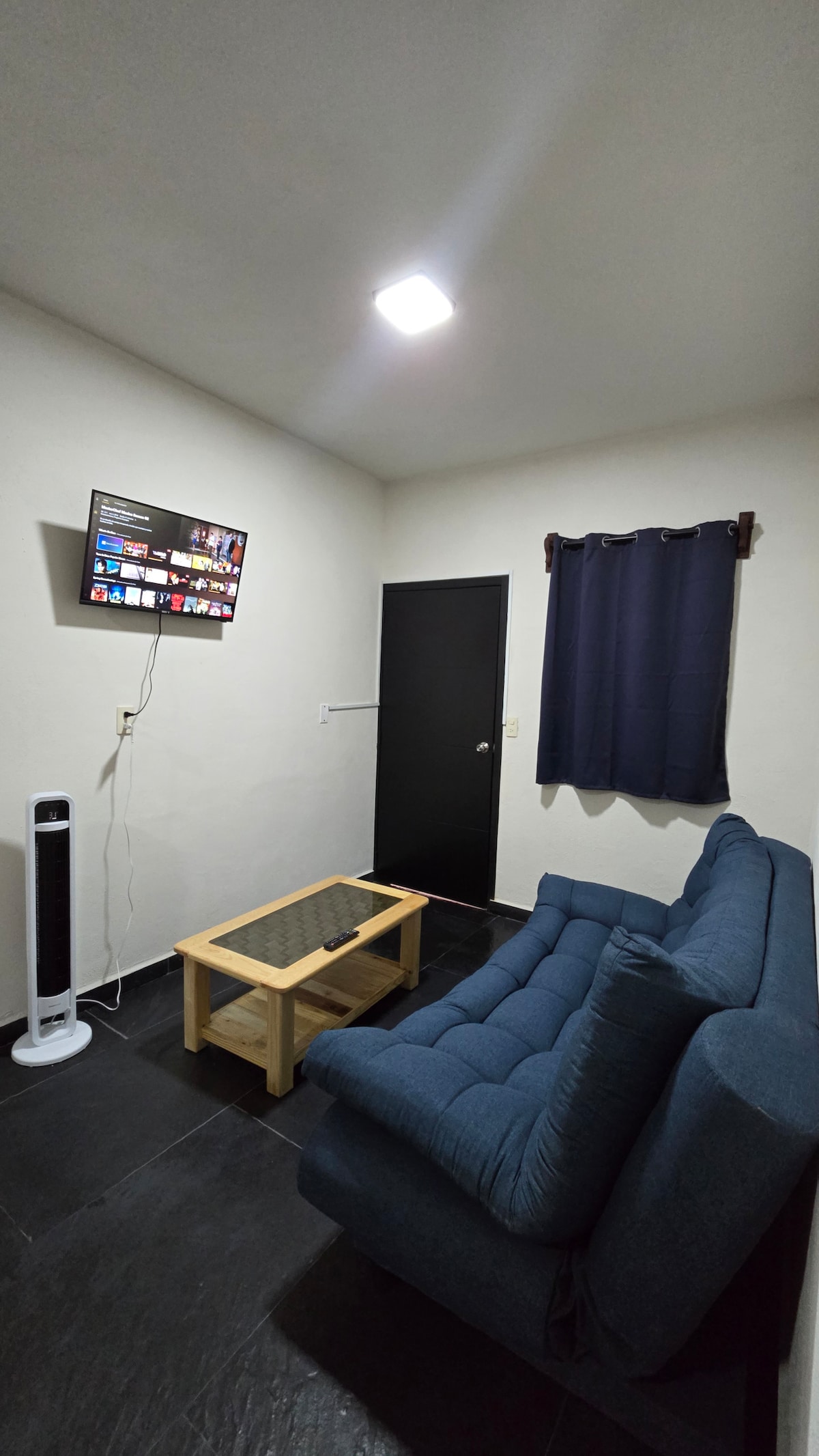 A modern living area features a blue sofa and a glass-top coffee table. A flat-screen television is mounted on the wall, displaying various content options. A dark curtain partially covers a window, and a doorway leads to another room.