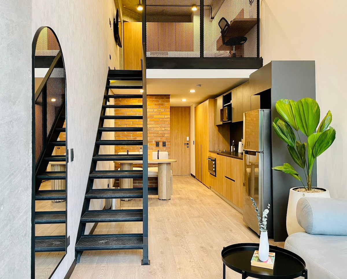 The image displays a modern loft interior highlighted by a sleek, black staircase leading to a lofted area. A fully equipped kitchen is visible, featuring wooden cabinetry and stainless steel appliances. A cozy living area is arranged with a comfortable sofa and decorative plants, enhancing the ambiance.