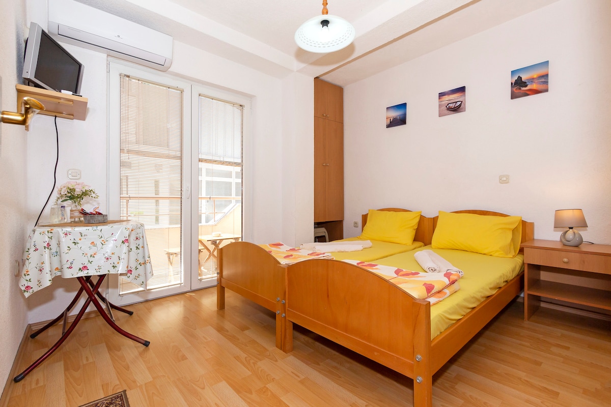 A well-lit sleeping area features a spacious bed with bright yellow bedding. A small table with a floral tablecloth is positioned near a large window, which opens to an outdoor balcony. Wall-mounted artwork enhances the neutral-colored walls, while a flat-screen TV and bedside lamp complete the space.