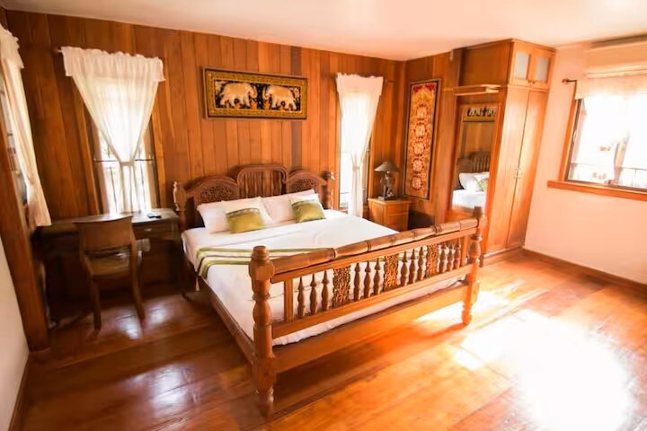 A spacious room features wooden walls, a king-size bed with decorative headboard, and two bedside lamps. A desk is positioned against one wall. Natural light filters through two windows, complemented by sheer white curtains. A wardrobe is situated in the corner, enhancing the room's storage options.