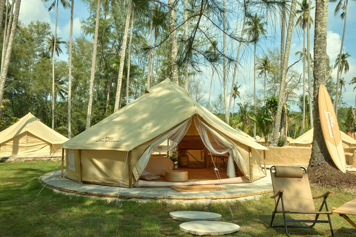 A spacious glamping tent is nestled among swaying palm trees, featuring a semi-transparent entrance that leads to an inviting interior. The tent's warm hues complement the surrounding greenery, creating a serene atmosphere. Lightweight seating is placed nearby, enhancing the outdoor experience.
