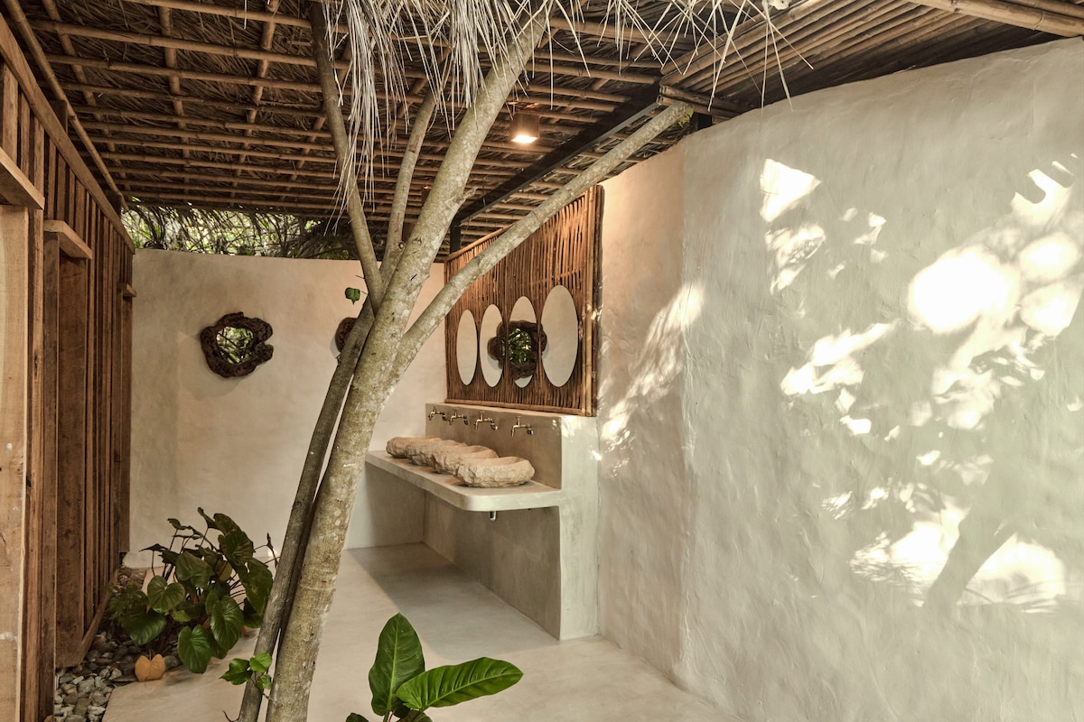 An outdoor shower area is featured, constructed with natural materials. Circular mirrors are mounted on a textured wall, while a stone base supports two cushioned benches. Lush green plants are arranged along the edges, with soft shadows created by a tree overhead.