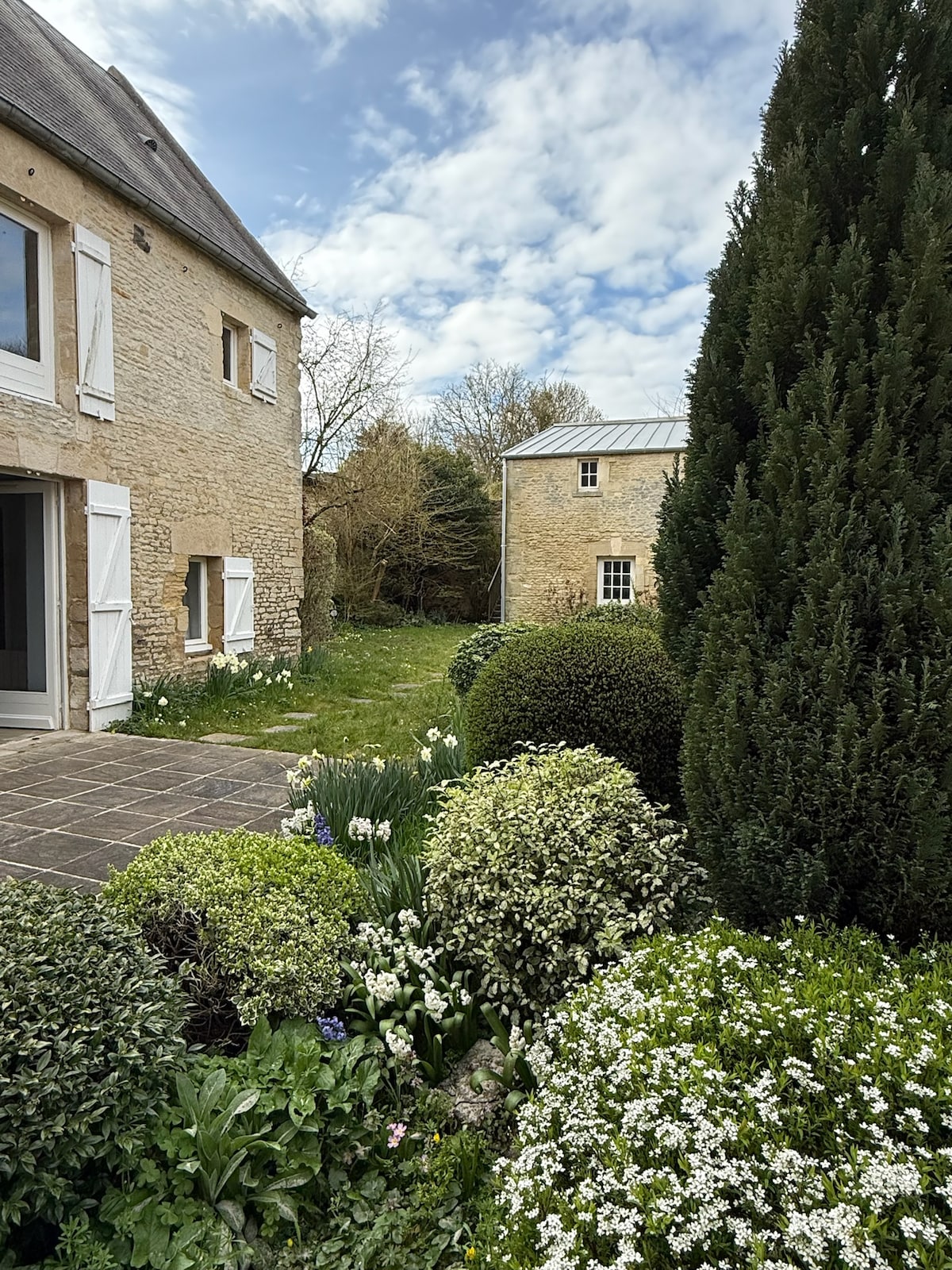 A serene outdoor area showcases a well-maintained garden featuring neatly trimmed hedges and blooming flowers. The stone facade of the house is complemented by light-colored shutters, and a second building is visible in the background, surrounded by a lush green landscape.