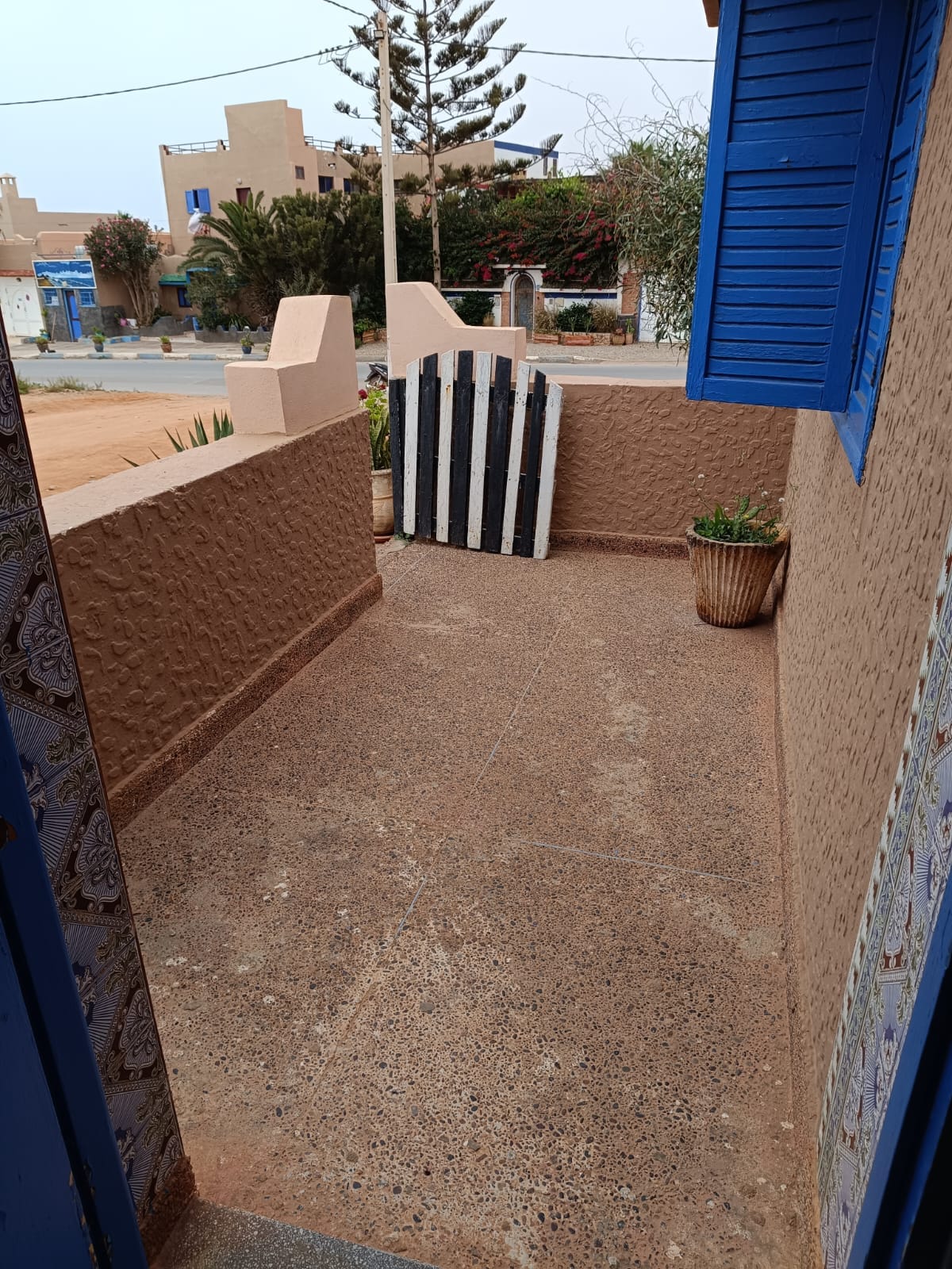 A small, outdoor terrace is visible, featuring a smooth pebble surface. The space is bordered by textured walls and includes a potted plant. A white wooden gate with black stripes stands to one side, while a glimpse of greenery can be seen beyond the terrace.