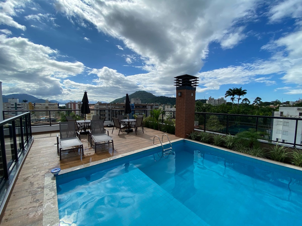 An outdoor leisure area features a swimming pool surrounded by wooden decking, lounge chairs, and tables with umbrellas. A picturesque view of the mountains and cityscape is visible under a partly cloudy sky.