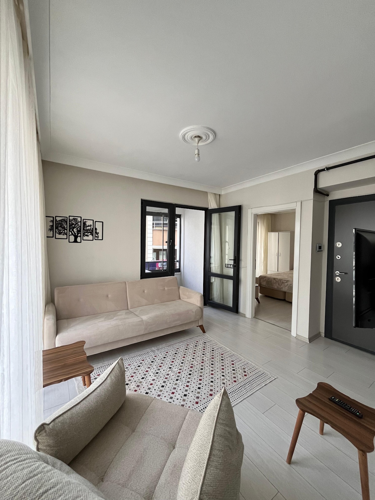 A light and airy living area is shown, featuring a comfortable sofa and an armchair set on a patterned rug. Large windows allow natural light to fill the space, with the entrance to a bedroom and additional doorway visible in the background.