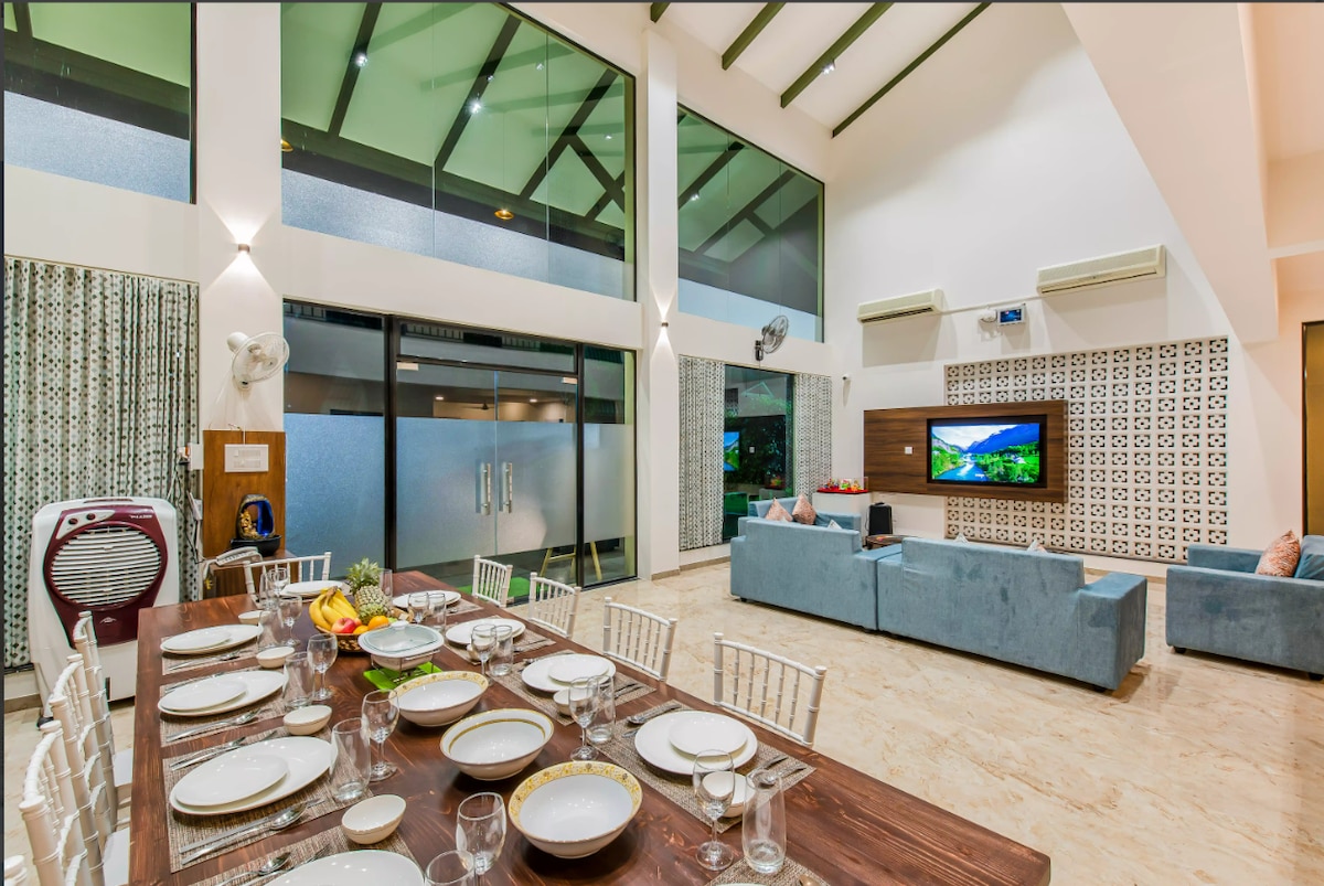 A spacious living area features a large dining table set for multiple guests, complemented by elegant dinnerware. Nearby, a cozy seating arrangement invites relaxation, while floor-to-ceiling windows allow natural light to fill the space. Decorative accents and a television complete the warm atmosphere.