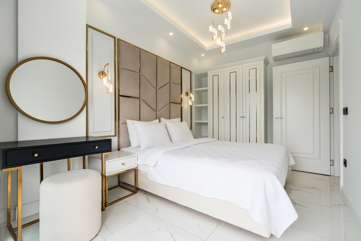 A modern bedroom showcases a soft color palette, featuring a spacious bed with crisp white linens. A large mirror is positioned beside a sleek vanity table. Elegant lighting fixtures and minimalistic storage cabinets enhance the room's contemporary design.