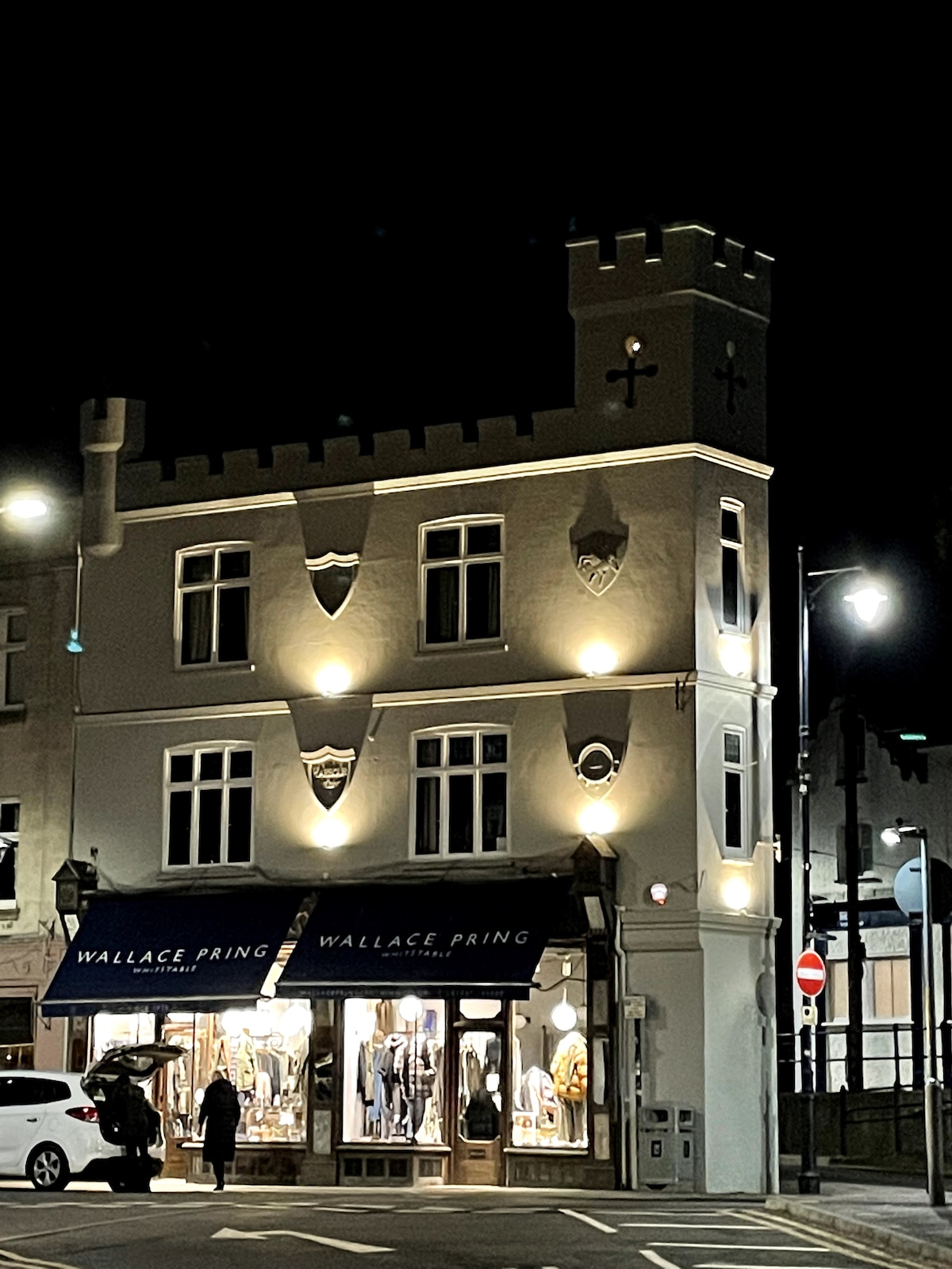 The building, illuminated at night, features a unique castle-like architecture with decorative shields and turrets. A clothing boutique, Wallace Pring, occupies the ground floor, with stylish displays visible through large windows. Streetlights cast a warm glow on the surrounding area.