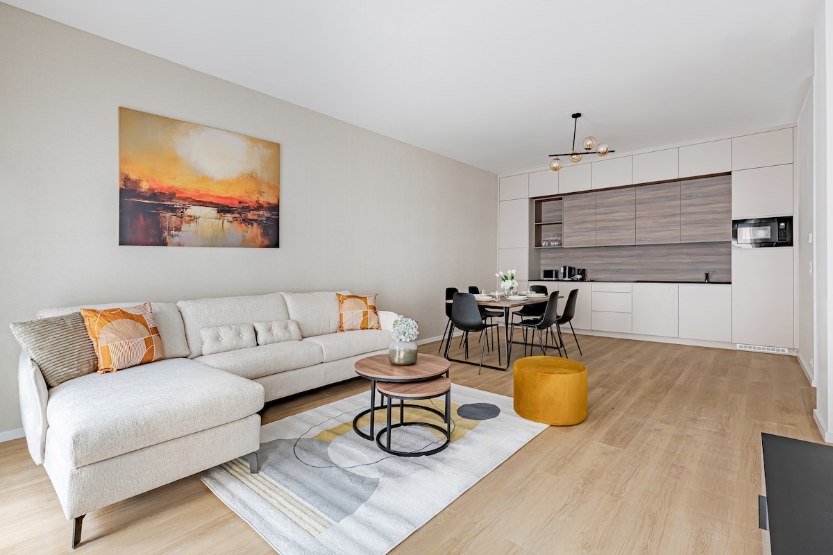 A modern living area features a light-colored sectional sofa and a round coffee table. A textured rug sits beneath, while a painting in warm tones is displayed on the wall. A dining set with four chairs is positioned near the kitchen area, which includes sleek cabinetry.