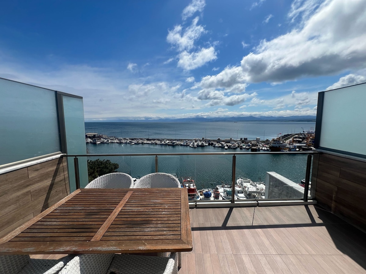 A spacious balcony with a wooden table and white chairs overlooks a scenic marina. The water reflects the sky's colors, with boats docked nearby, framed by distant mountains under a partly cloudy sky.