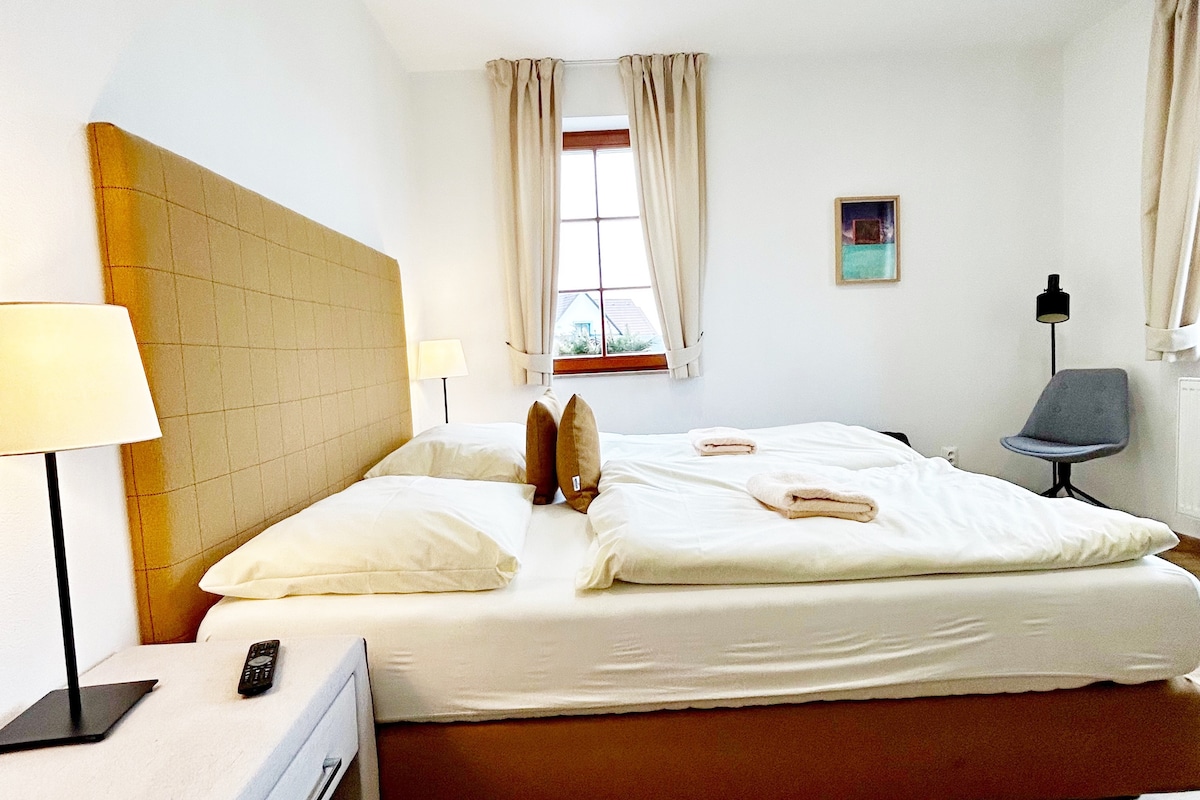 A well-lit bedroom features a spacious bed dressed in crisp white linens. Pillows are neatly arranged, and bedside lamps provide soft illumination. A window with light curtains allows natural light to enter, while a minimalist chair and a small nightstand complete the serene atmosphere.