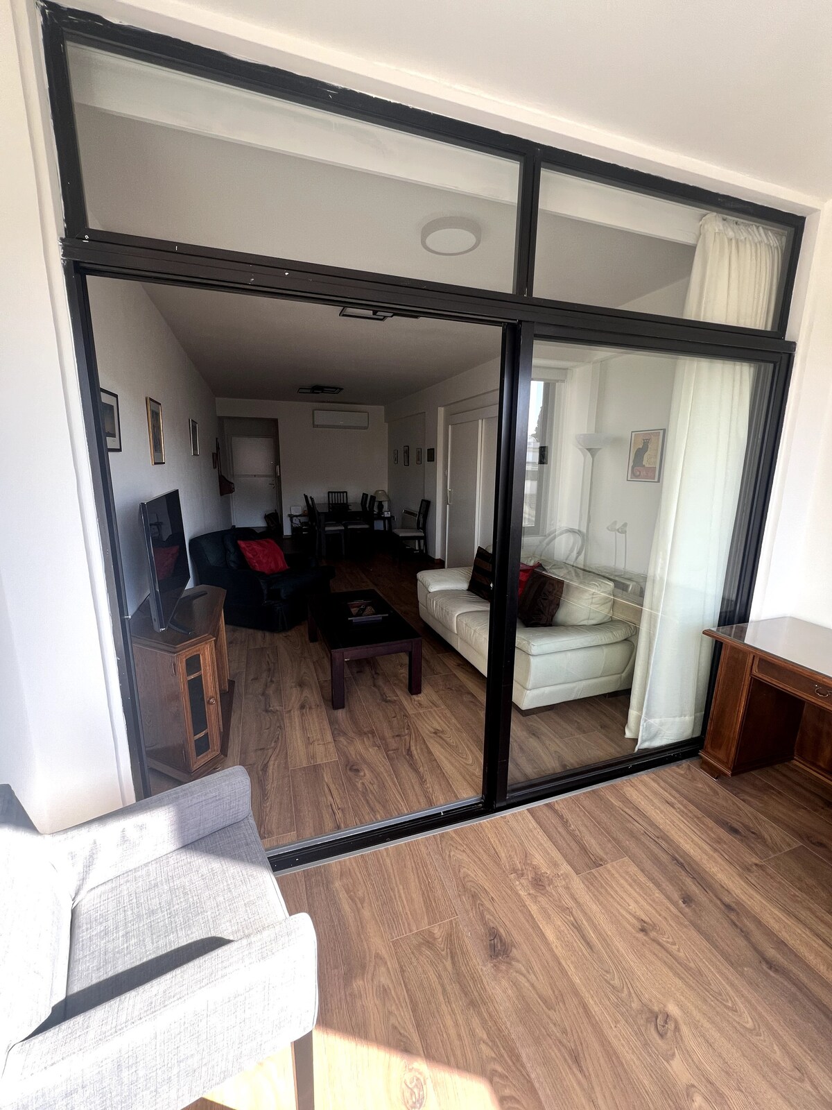 A spacious living area is visible through large sliding glass doors that allow abundant natural light. The room features a light-colored sofa and dark wooden furniture. A flat-screen TV is placed on a console, while dining chairs and a table can be seen in the background.