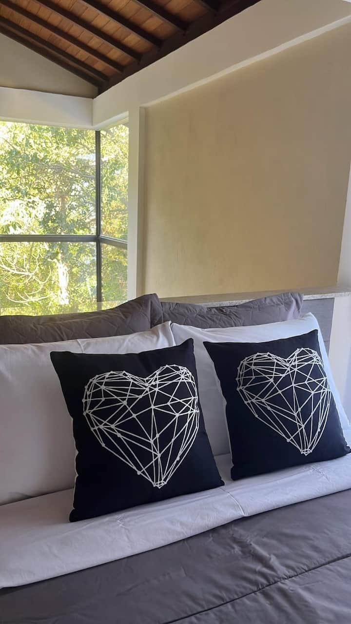 Black pillows with a geometric heart print on a bed with gray and white sheets. In the background, a light beige wall and a window overlooking a green area, bringing natural light to the cozy and modern environment.