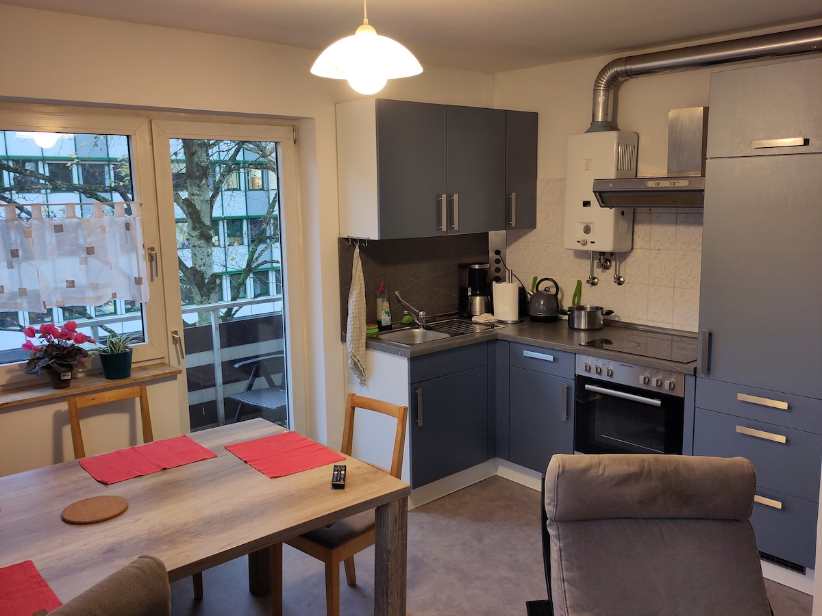 A modern kitchen with dark cabinetry and stainless steel appliances is visible. A dining table with red placemats is placed centrally, complemented by wooden chairs. Natural light enters through large windows, revealing a small outdoor area adorned with plants.