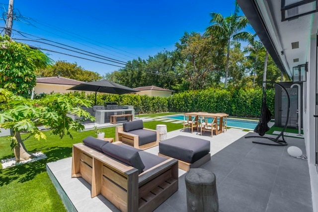 An outdoor lounge area features wooden seating with dark cushions, complemented by a dining table set for several guests. Nearby, a private pool is surrounded by lush greenery and palm trees, while a shaded area with a large umbrella offers respite from the sun.